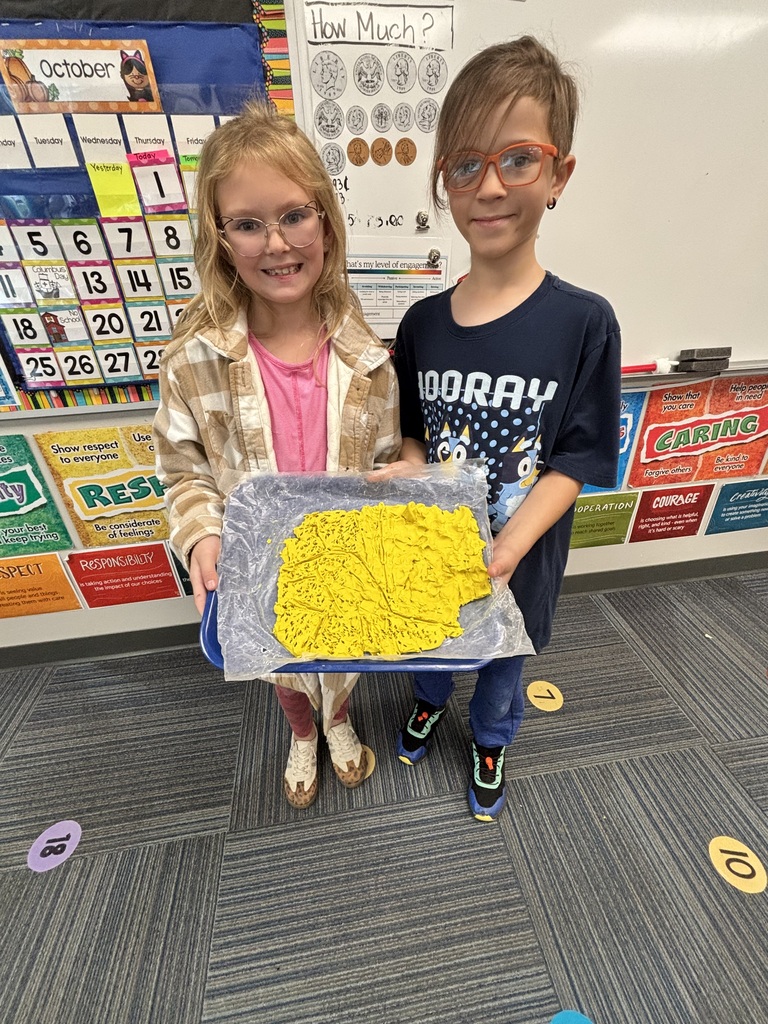 Two students standing together holding up a tray filled with textured yellow clay in front of classroom learning charts.