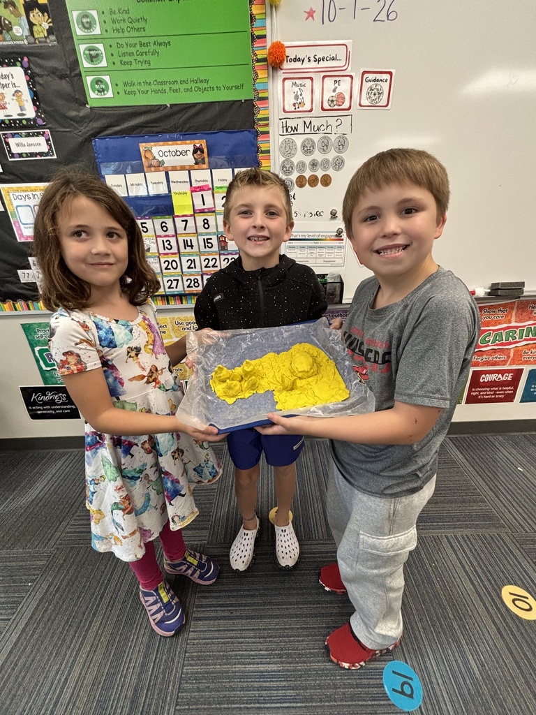 A group of three young students posing with their yellow dough sculpture on a clear-wrapped tray.