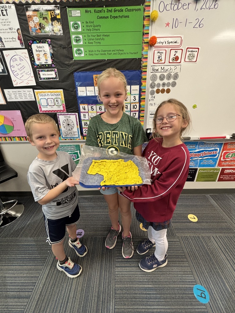 Three smiling classmates standing in a line presenting their yellow play dough project on a blue tray.