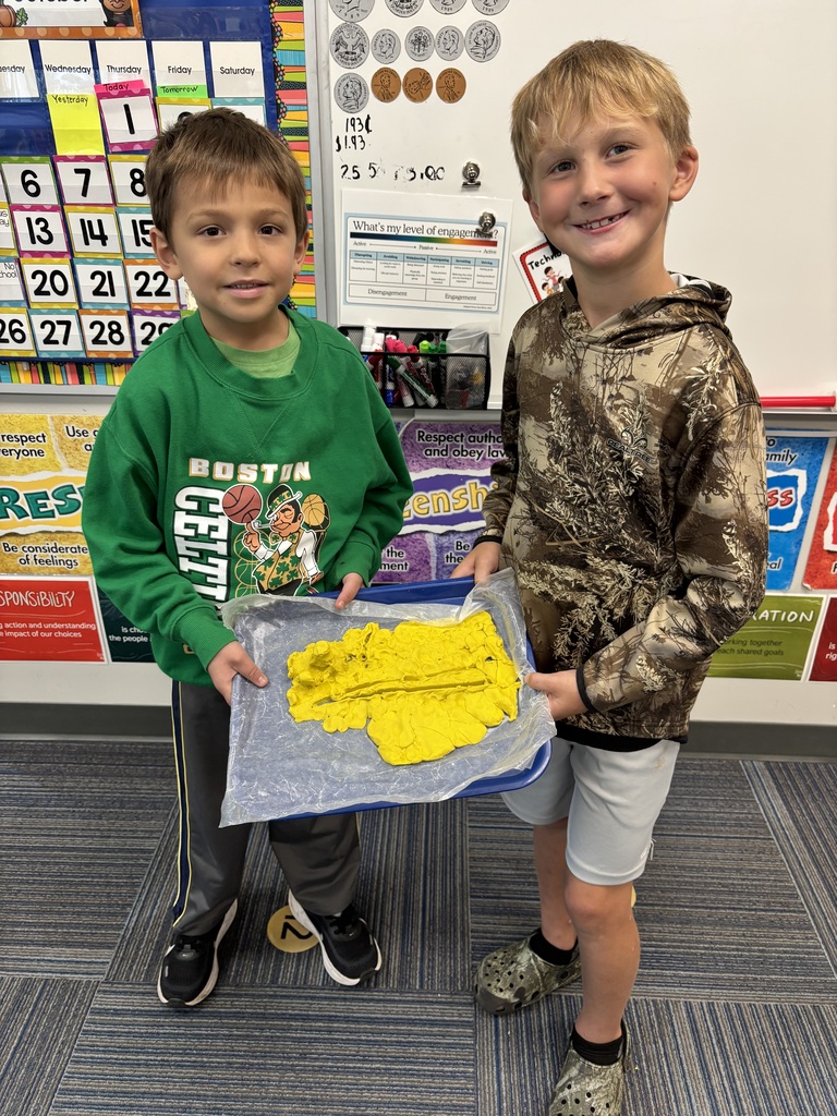 Two boys smiling while holding up a blue tray displaying their finished yellow clay sculpture in front of a classroom bulletin board.