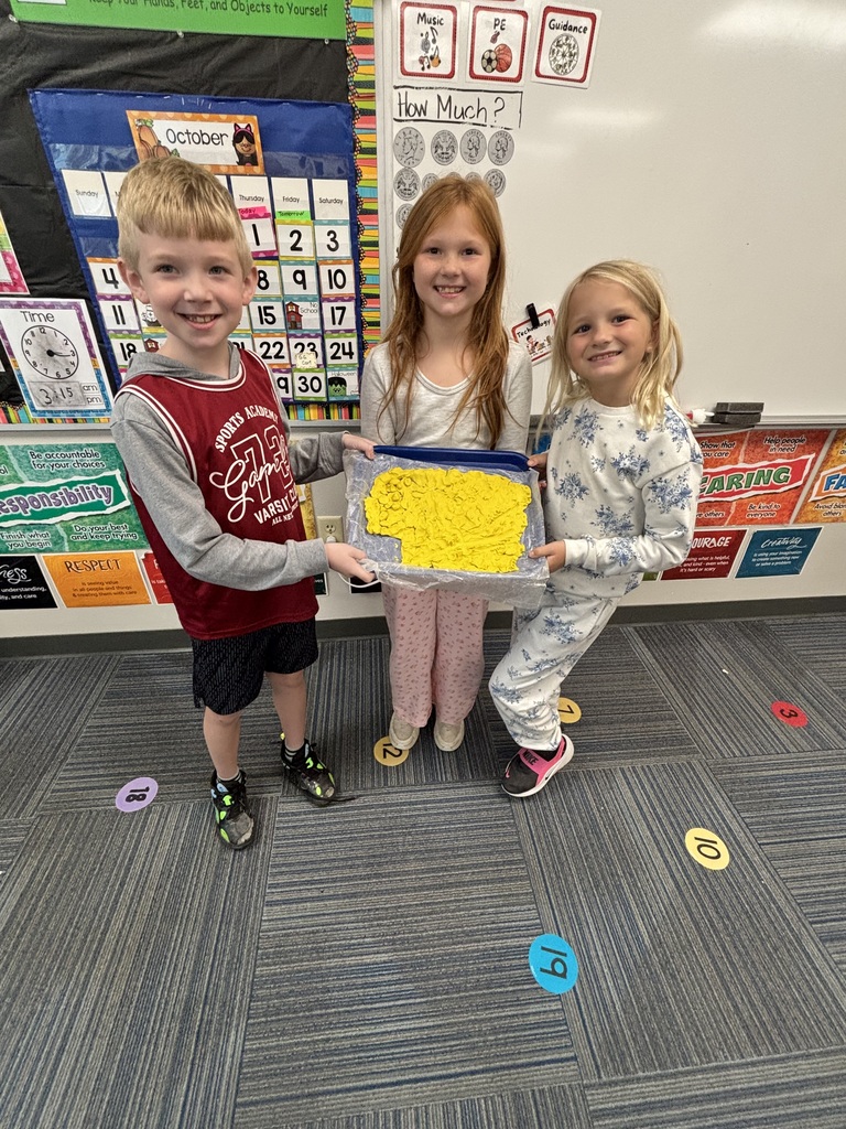 Three students smiling for the camera while holding a blue tray with a large yellow clay shape in their classroom.