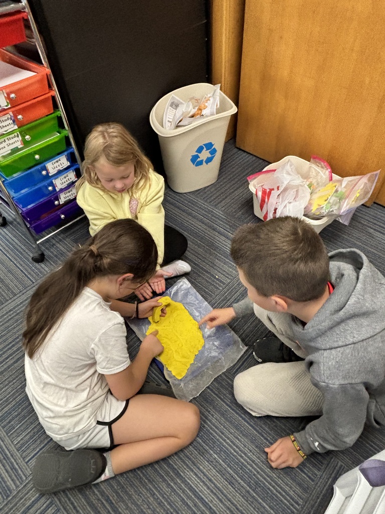 A group of three young students kneeling on the floor to work collaboratively on a yellow clay project.