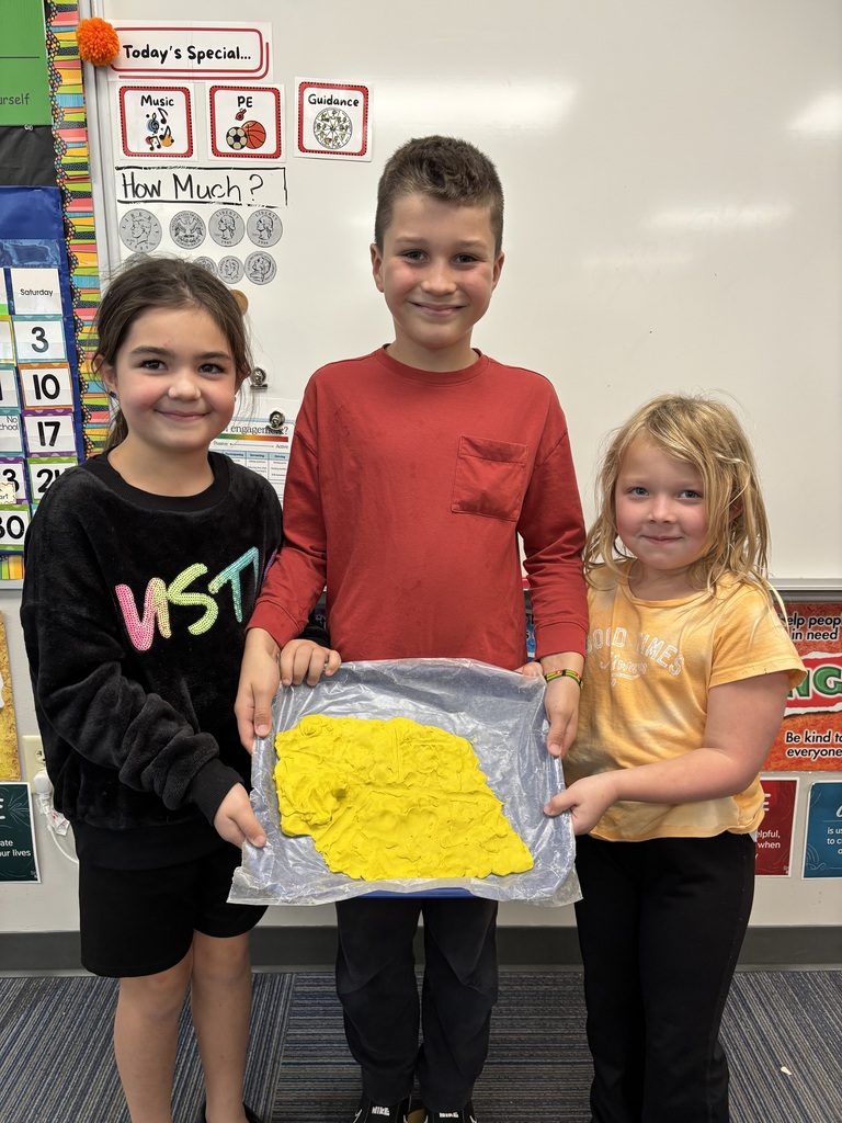 Three elementary students posing proudly together behind a blue tray holding their flattened yellow modeling dough creation.