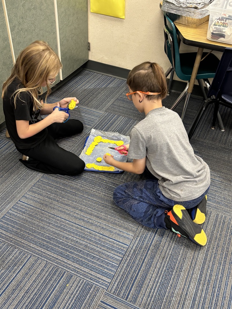 Two students kneeling on the classroom floor while using scissors to shape yellow modeling clay on a plastic-covered tray.