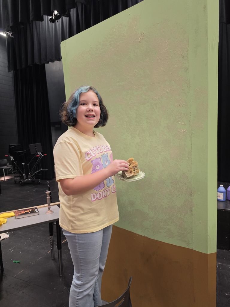 A girl is smiling at the camera in front of a green wall painting it. 