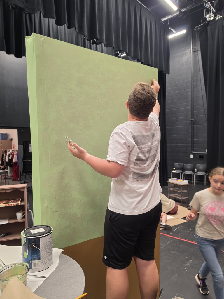 A boy has his back to the camera and is painting a green wall for set design. 
