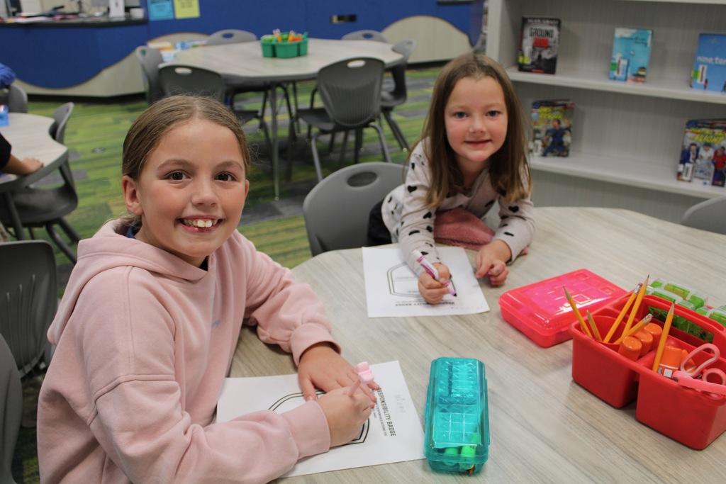 A fifth and first grade girl sit at a table working on a responsibility shield.