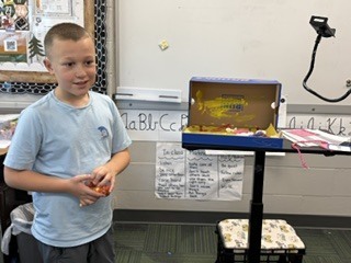 A boy standing near a presentation stand and talking about his handcrafted shoebox diorama.