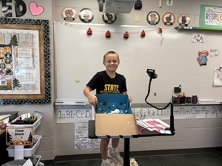 A boy wearing a black "State" t-shirt smiling proudly behind his shoebox ecosystem diorama.