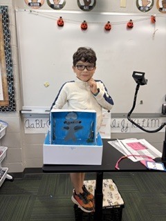 A student wearing glasses posing next to his blue-themed diorama project set on a classroom presentation cart.