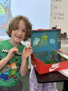 A student in a green Minecraft shirt smiling while displaying a shoebox diorama project and holding a small figure.