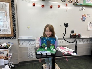 A young girl standing behind a podium presenting her detailed, colorful diorama built inside a box.