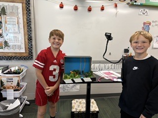 Two boys standing side by side in front of a whiteboard presenting a nature diorama created inside a cardboard box.