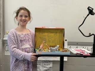 A smiling girl standing beside her safari-themed shoebox diorama filled with toy animals.