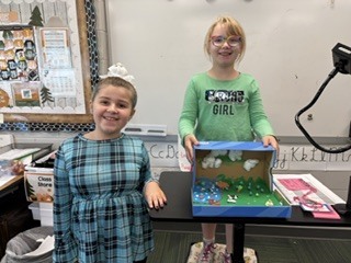 Two smiling students standing together at a presentation cart showing off their blue sky and green landscape diorama.