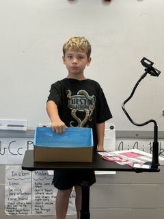 A student in a black t-shirt standing at a classroom presentation stand with his blue shoebox diorama.