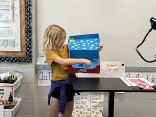 A student in a yellow shirt pointing to figures inside her open Nike shoebox diorama during a presentation.
