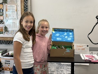 Two girls posing happily together next to a open cardboard diorama displaying a cloudy sky lid.