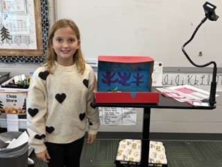 A young girl wearing a cream-colored sweater with black hearts smiling beside her red shoebox project.