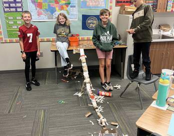 Four students pose near their construction of a model roller coaster. One student stands on a chair. The coaster is made of paper towel tubes and tape.