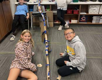 Four students pose next to a construction of a model roller coaster made of paper towel tubing and tape.