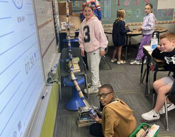 Five students stand and crouch as they work on their model roller coaster. It stands about four and a half feet tall and is made with paper towel tubing and tape. 