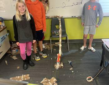 Three students stand next to a model roller coaster constructed by paper towel tubing and tape. They look happy!