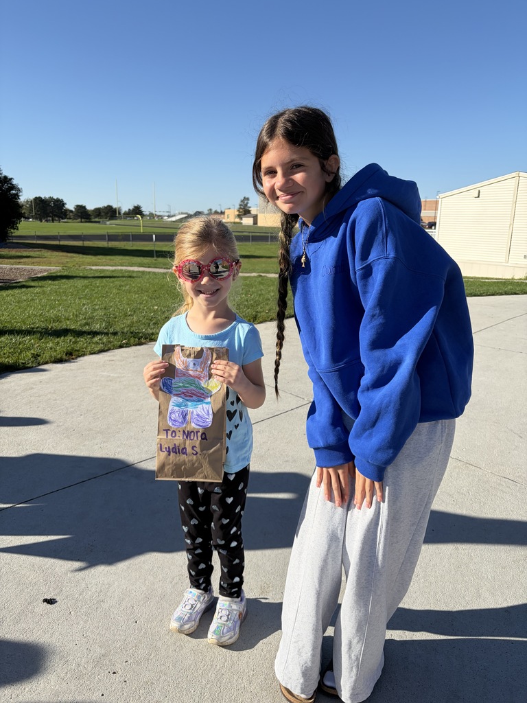A young girl wearing heart-patterned leggings holding a bag labeled "To: Nora / Lydia S." next to a girl in a royal blue hoodie.