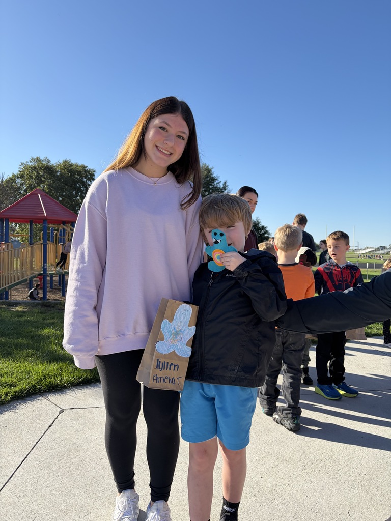 An older girl in a light purple sweatshirt and a younger boy in blue shorts holding a blue cutout character and a paper bag labeled "Tyllen / Amelia T".