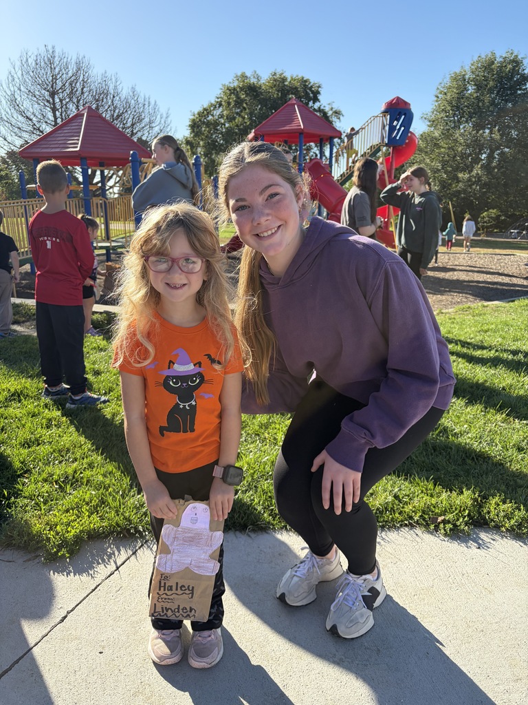 A young girl with blonde hair and glasses in an orange cat shirt holding a bag decorated with a gingerbread man, standing next to an older girl in a dark purple hoodie.