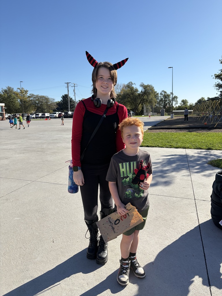An older student with red/black striped horn headband and headphones posing next to a red-haired boy holding a Hulk t-shirt and a brown paper bag.
