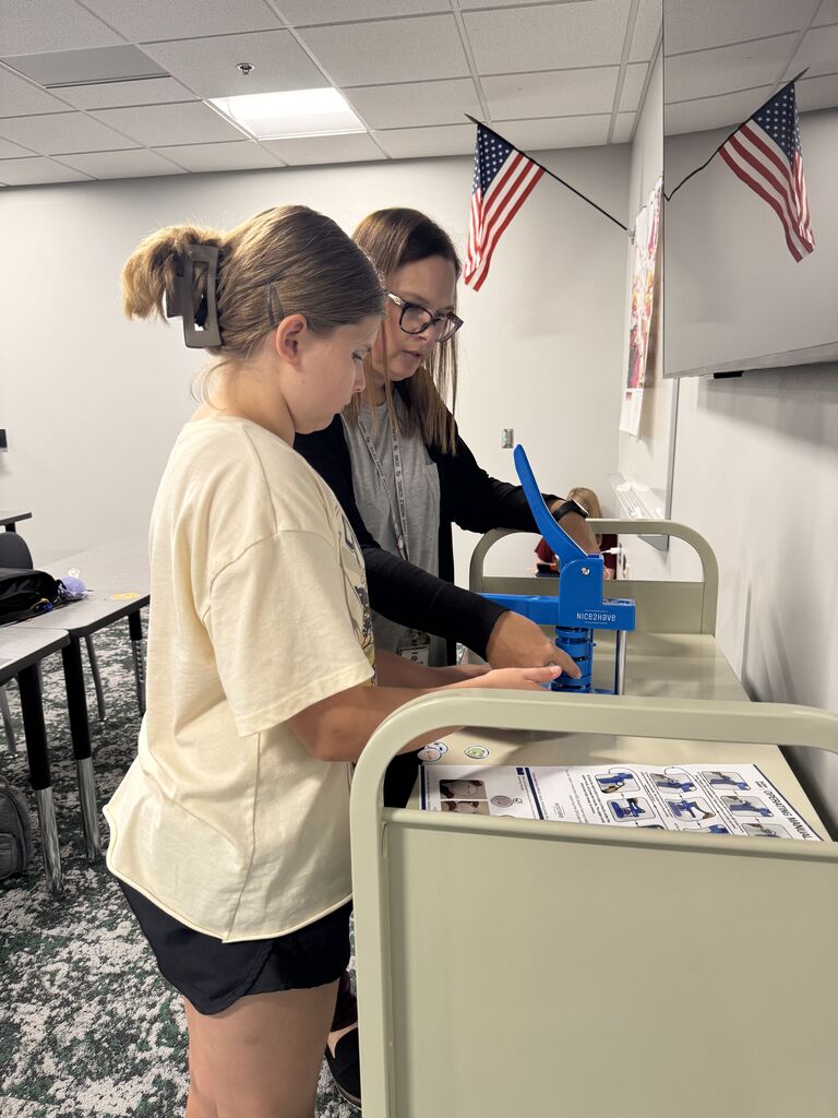 A female student stands next to a female teacher and is using a blue button maker machine. 