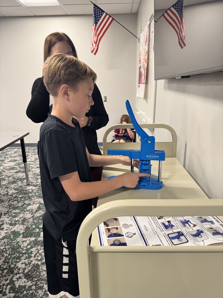 A male student stands in front of a blue button making machine lining up his items. 
