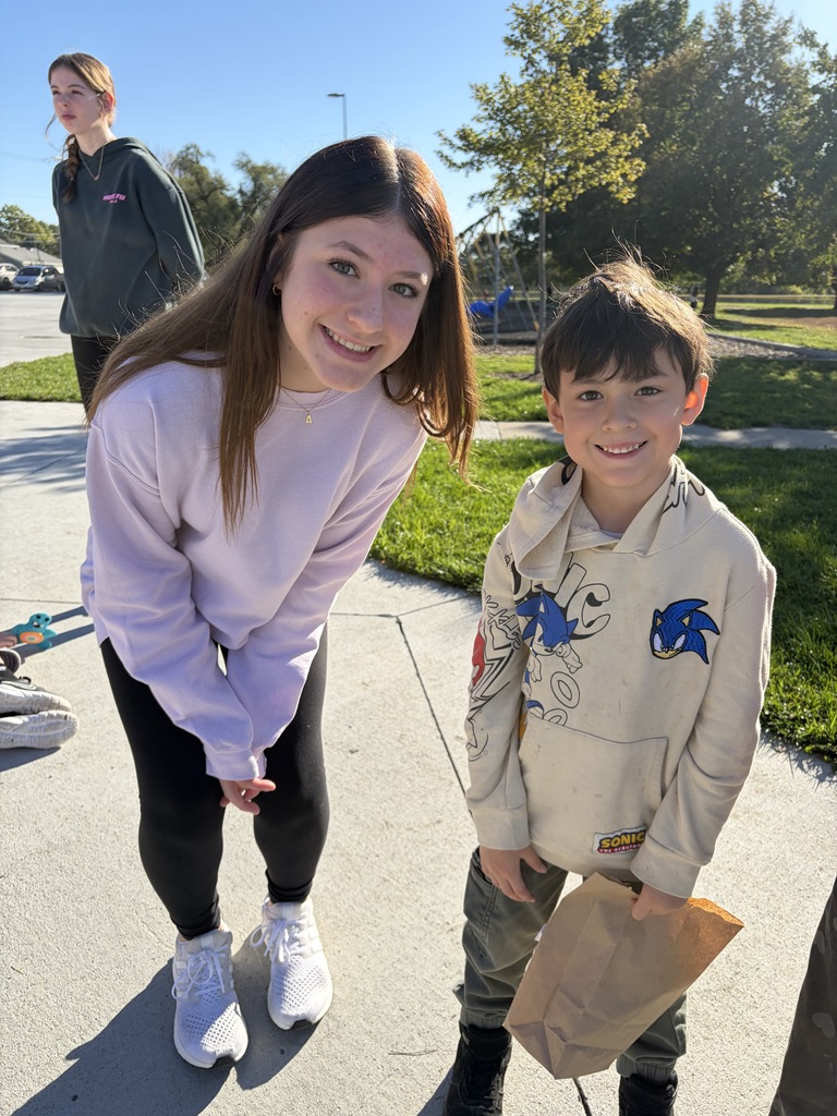 A young woman in a light pink/purple sweatshirt kneeling beside a young boy wearing a cream-colored Sonic the Hedgehog hoodie, who is holding a brown paper bag on a concrete walkway.