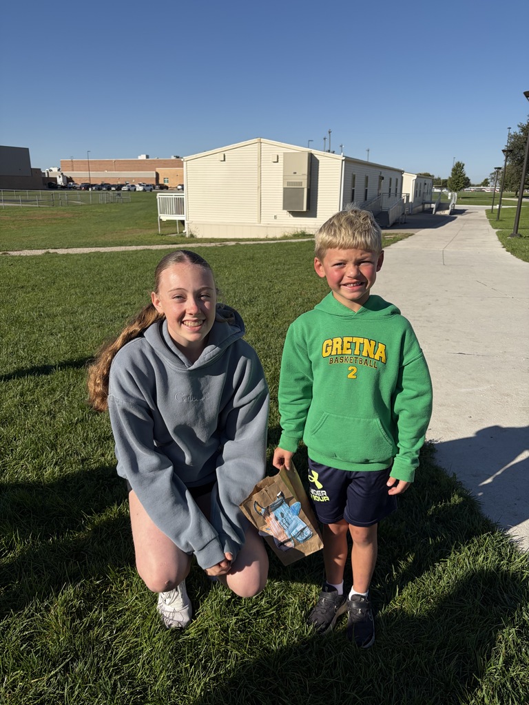 A young woman crouching down in a blue/grey hoodie next to a young boy wearing a green "Gretna Basketball 2" hoodie, who is holding a paper bag with a blue monster drawing on it.