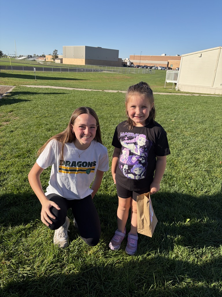 A young woman crouching in a white shirt that says "DRAGONS" next to a young girl wearing a black t-shirt and holding a paper bag on a lawn.