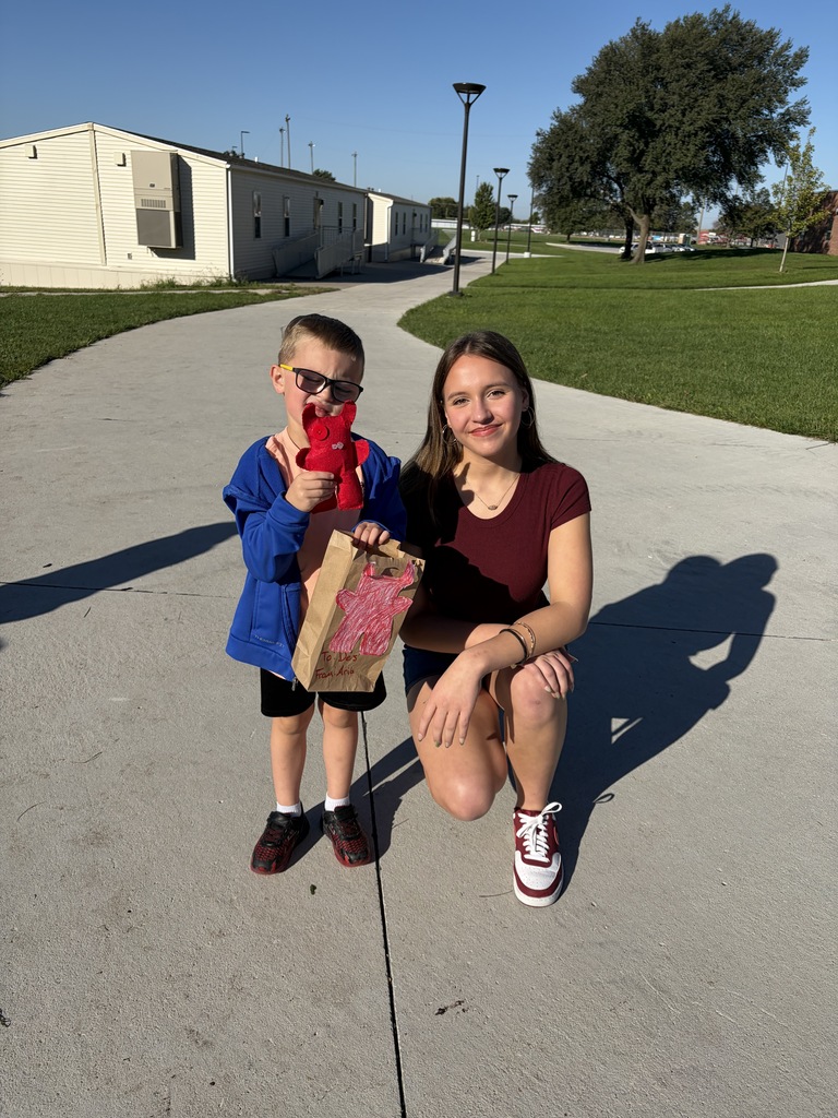 A young woman kneeling in a dark red top next to a young boy with glasses who is holding a red plush monster and a brown paper bag with a matching red monster drawing on it.