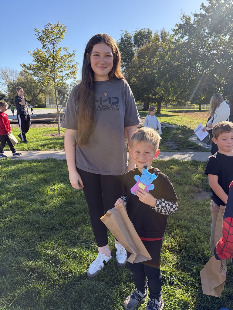 A young woman in a grey shirt standing beside a young boy in a black hoodie with checkerboard sleeves, who is holding up a light blue monster cut-out and a brown paper bag.