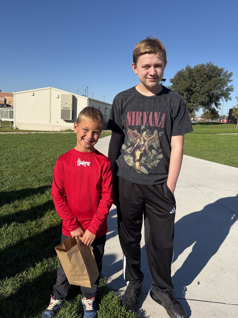An older boy wearing a black Nirvana t-shirt standing beside a younger boy wearing a red long-sleeve shirt (labeled "Huskers") holding a brown paper bag on a grassy field.