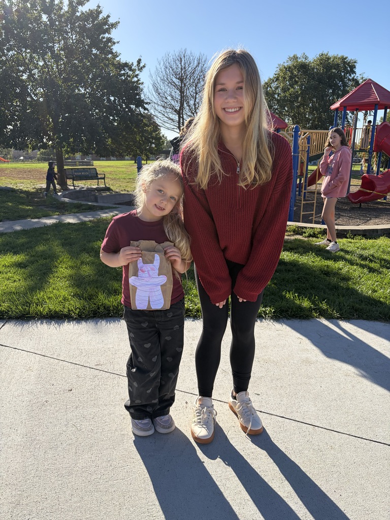 A young woman with long blonde hair in a dark red quarter-zip sweater standing next to a young girl wearing a maroon shirt, who is holding up a paper bag decorated with a purple monster drawing in front of a playground.