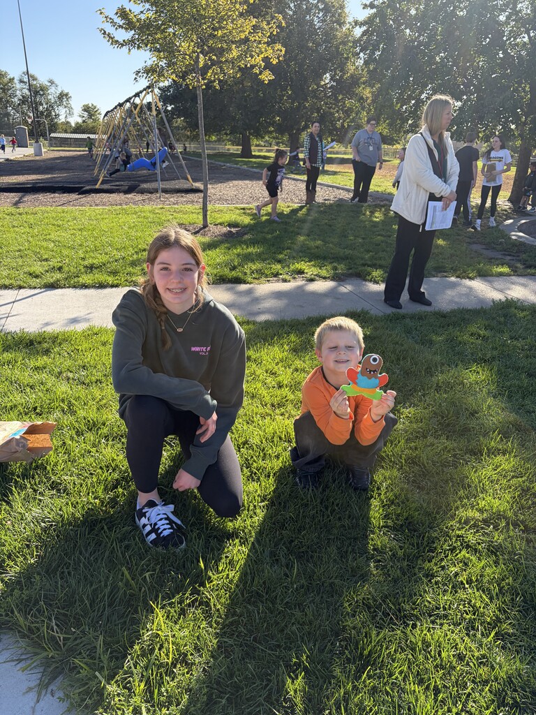 A young woman in a dark grey sweatshirt crouching in the grass next to a young boy wearing an orange shirt, who is smiling while holding up a colorful monster paper cutout.