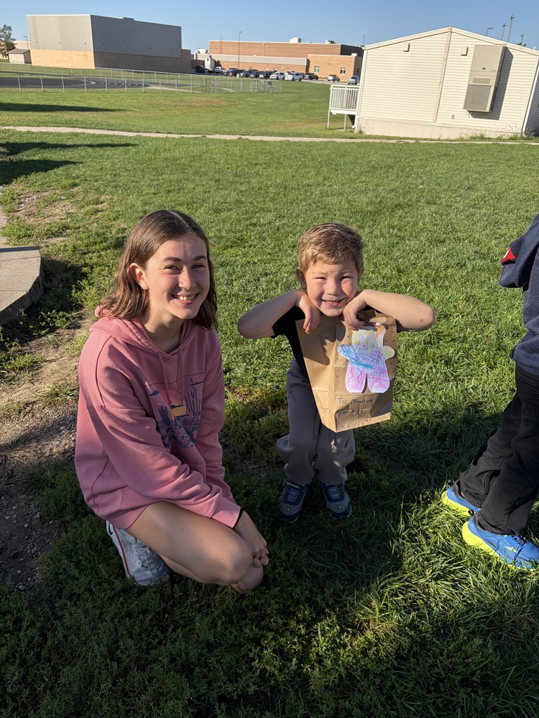 A young woman crouching in a pink hoodie next to a young boy who is proudly displaying a paper bag decorated with a colorful monster craft cutout on top.