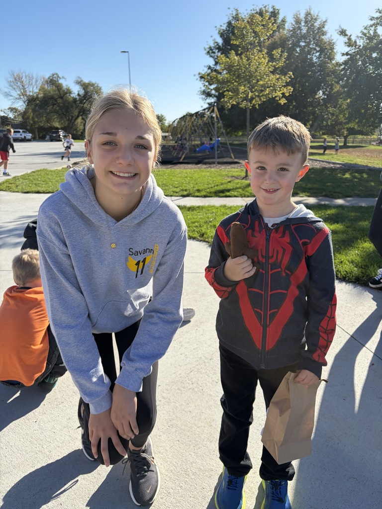 A young woman wearing a grey hoodie (labeled "Savannah #11") kneeling next to a young boy wearing a black-and-red Spider-Man zip-up hoodie, who is holding a stuffed brown object and a paper bag outdoors.