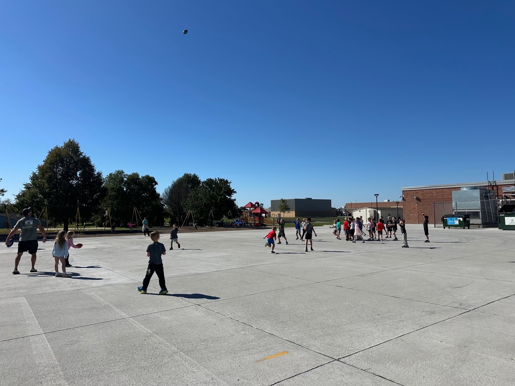 Children and an adult walking across a concrete schoolyard during an outdoor recess or football activity, with parked cars visible in the background.