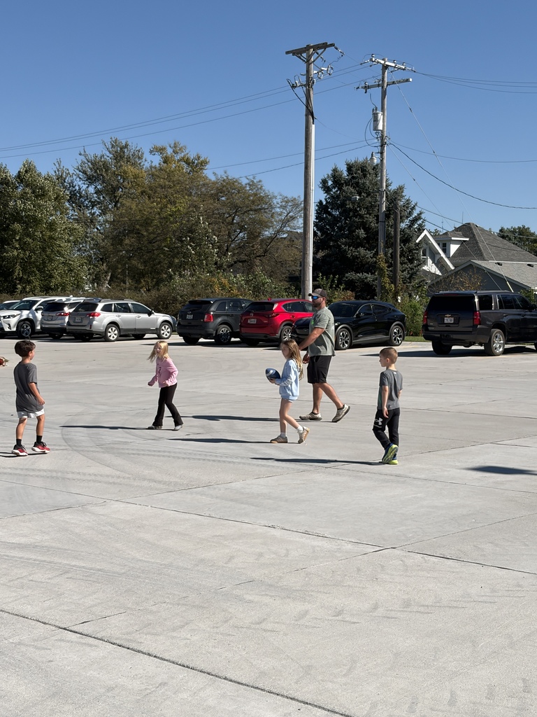 An adult throwing a football while kids play on the concrete blacktop outside a school building.