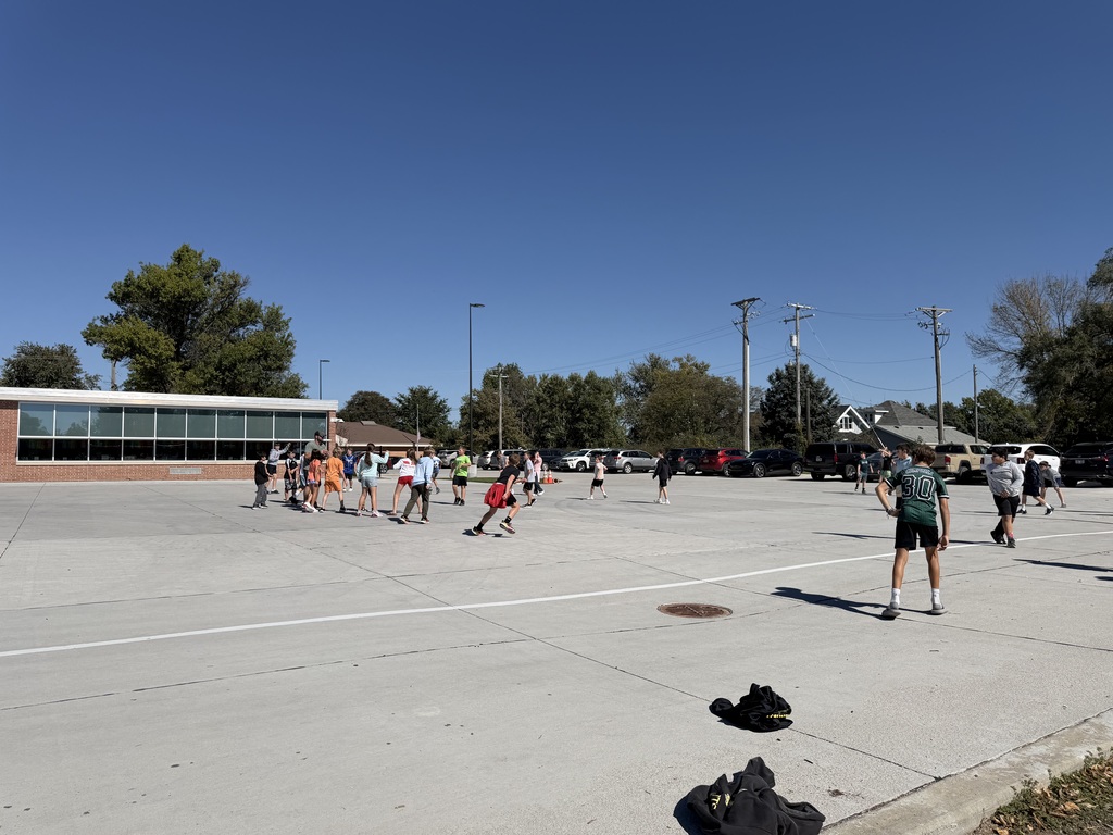 A wide perspective of kids running around playing football on a wide concrete field outside school buildings.