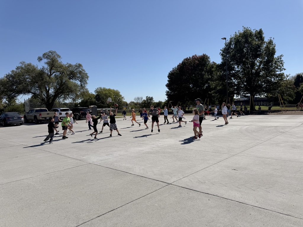 A large group of elementary school children playing a game of football on a sunny concrete playground.