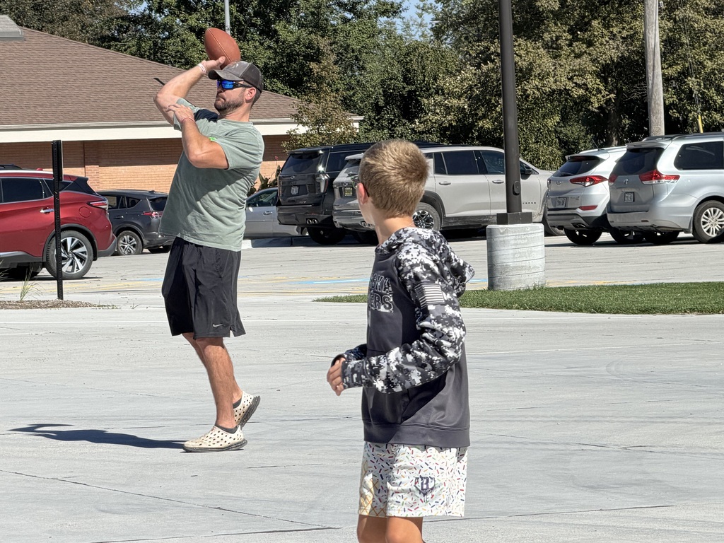 An adult in a cap and sunglasses winding up to throw a football while a young boy looks on from the foreground on a sunny day.