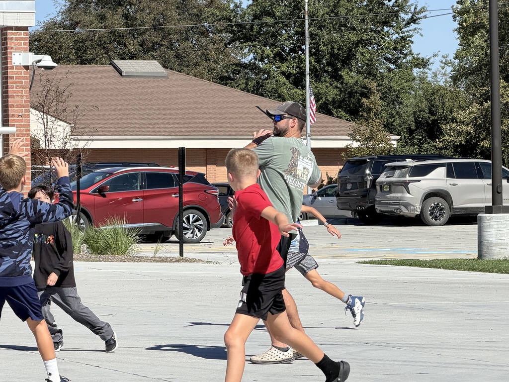 An adult wearing a cap and green shirt preparing to throw a football while kids run past him on the concrete playground.