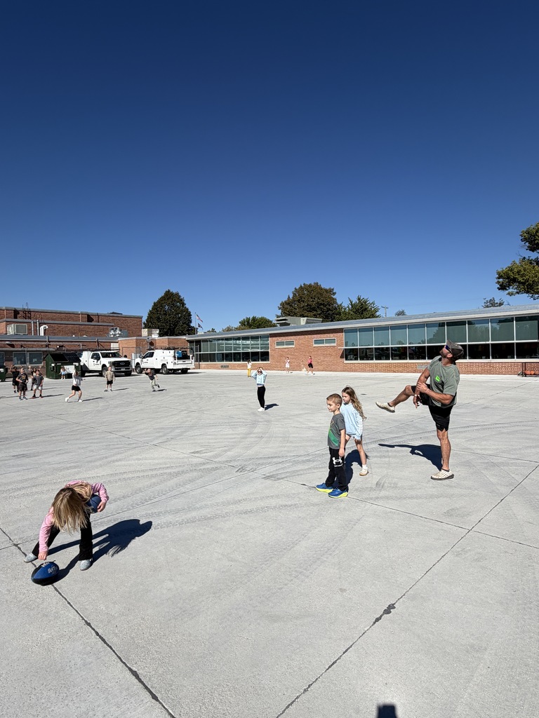 A wide shot of children playing an outdoor football game on a large concrete school play area under a clear blue sky.